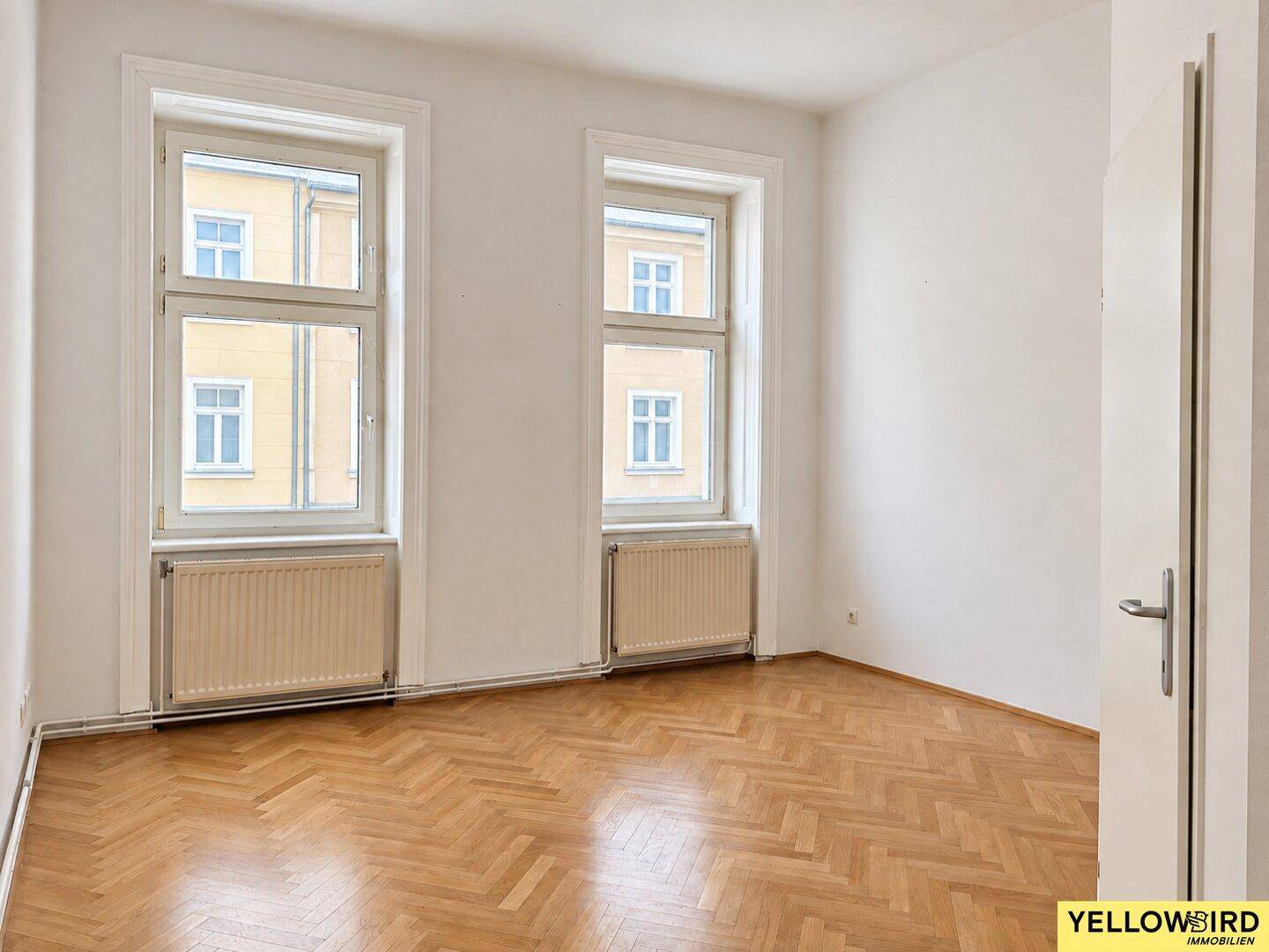 Heller Raum mit Fischgrätparkett und zwei Fenstern, ideal als Wohn- oder Schlafzimmer.