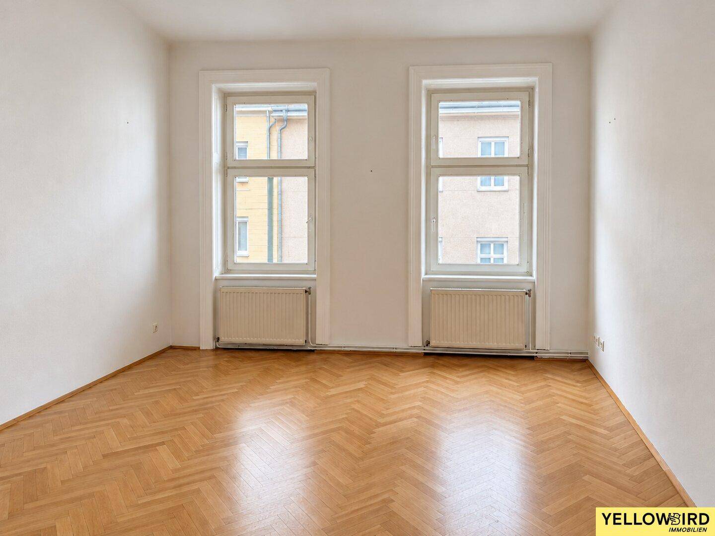 Geräumiges Zimmer mit zwei großen Fenstern und klassischem Fischgrätparkettboden.