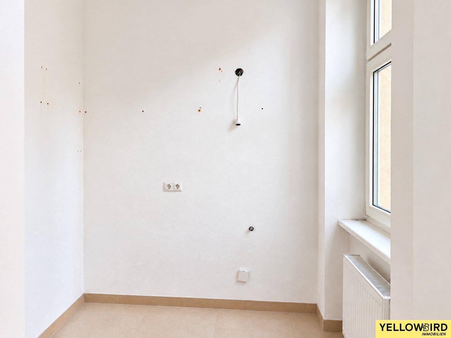 Leerer Küchenraum mit Fliesenboden, Fenster und Anschlüssen für eine Einbauküche.