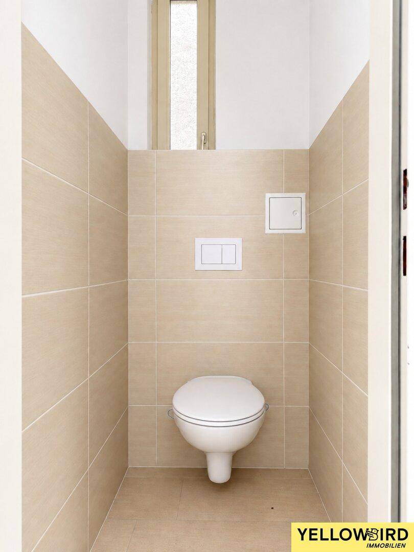 Separates WC mit beigefarbenen Fliesen, wandhängender Toilette und schmalem Fenster.