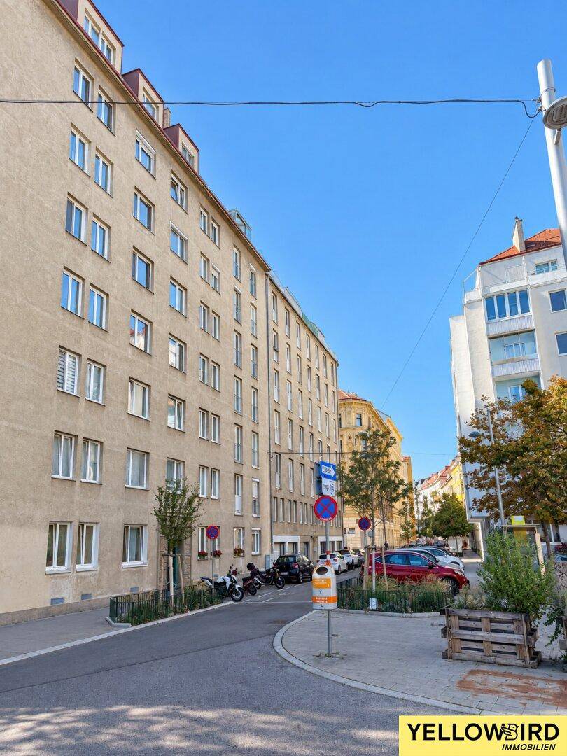 Mehrstöckiges Wohngebäude an einer städtischen Straße mit geparkten Autos und Bäumen unter blauem Himmel.