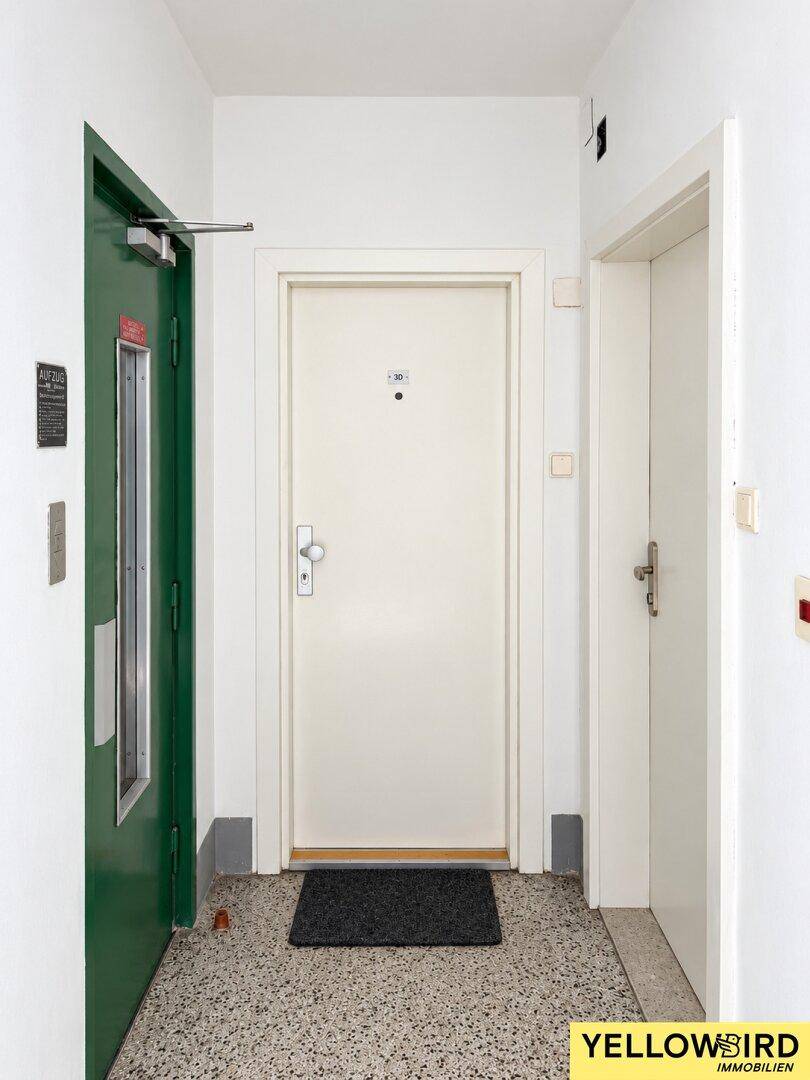 Heller Flur mit Terrazzoboden, weißer Wohnungstür und Zugang zu einem Aufzug.