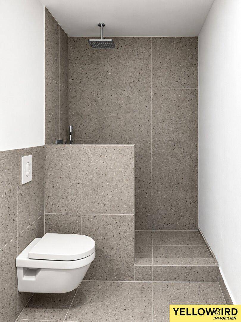 Modernes Badezimmer mit grauen Fliesen, wandhängendem WC und einer geräumigen begehbaren Dusche mit Regenduschkopf.