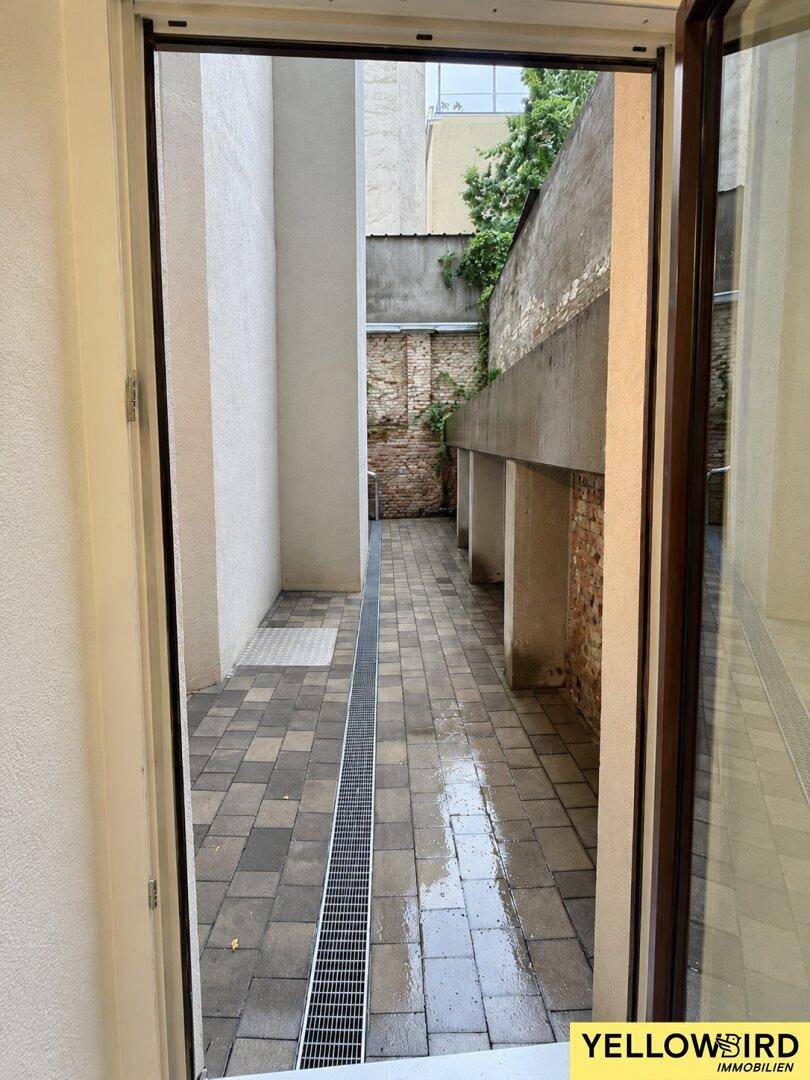 Blick von innen durch eine Tür auf einen schmalen, gepflasterten Innenhof mit Entwässerungsrinne.