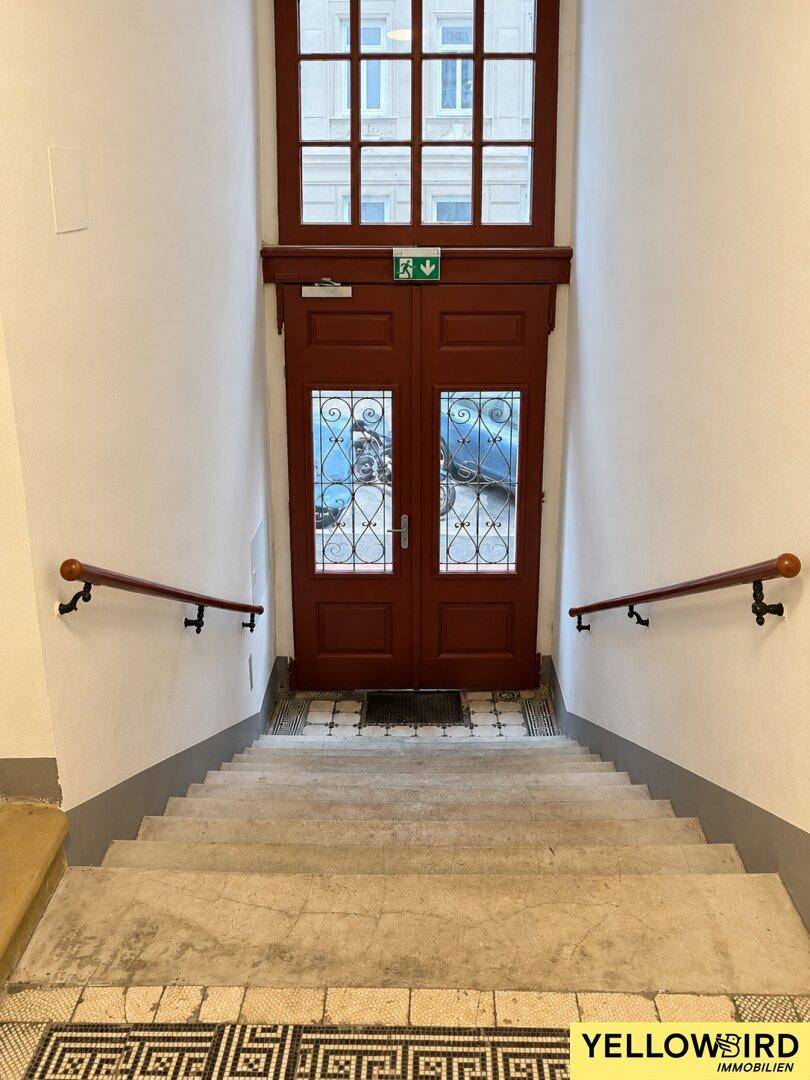 Historisches Treppenhaus mit breiten Stufen, Holzgeländern und einer großen Doppeltür mit Glasornamenten.