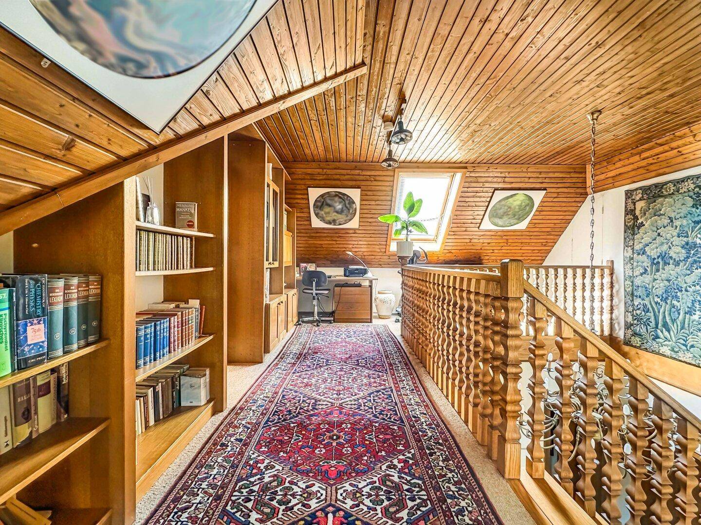 Helle Galerie mit Dachschrägen, Holzvertäfelung, Bücherregalen und Arbeitsbereich am Fenster.