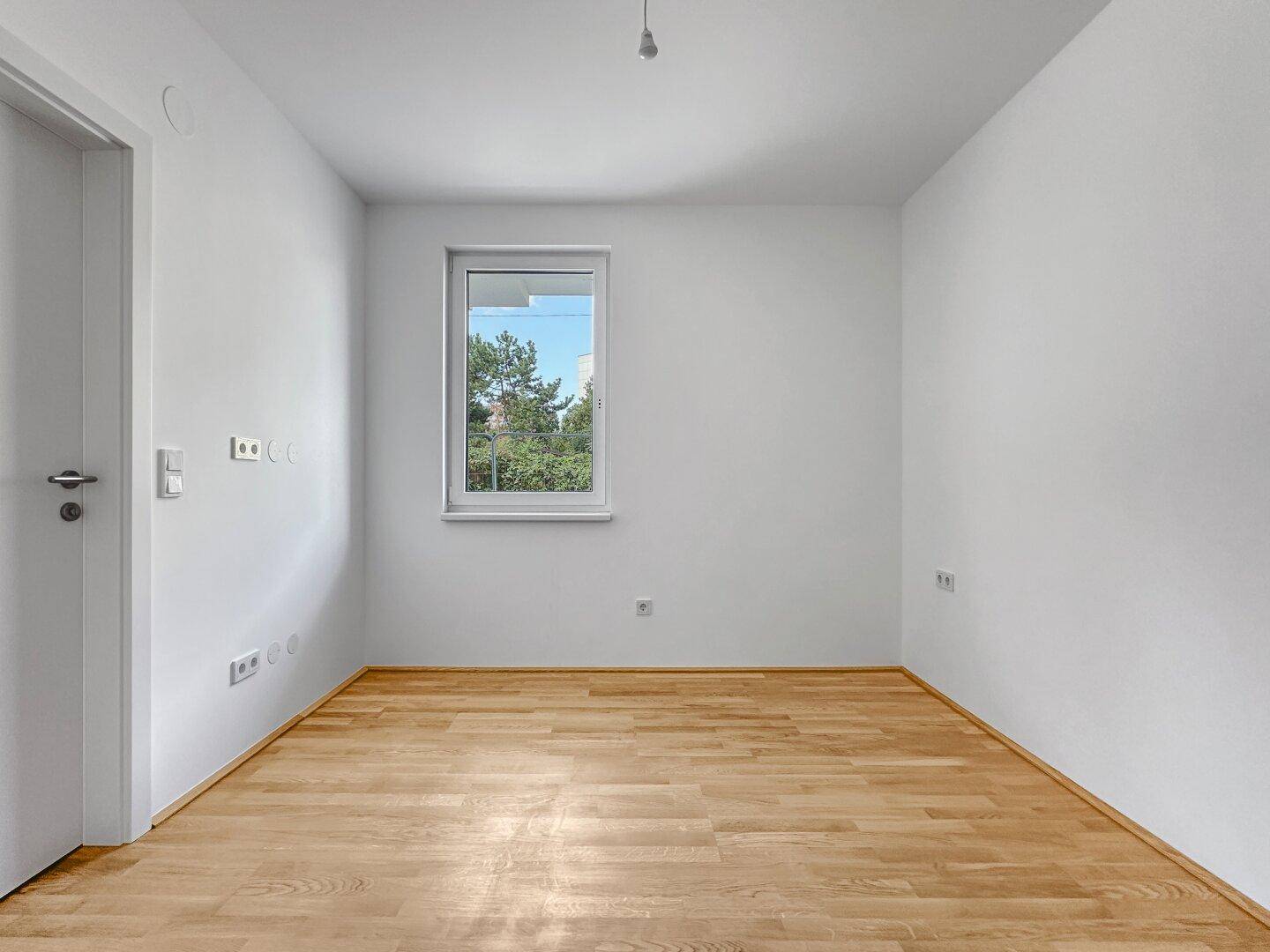 Ruhiges Zimmer mit hellem Parkettboden und einem Fenster mit Blick auf Bäume.