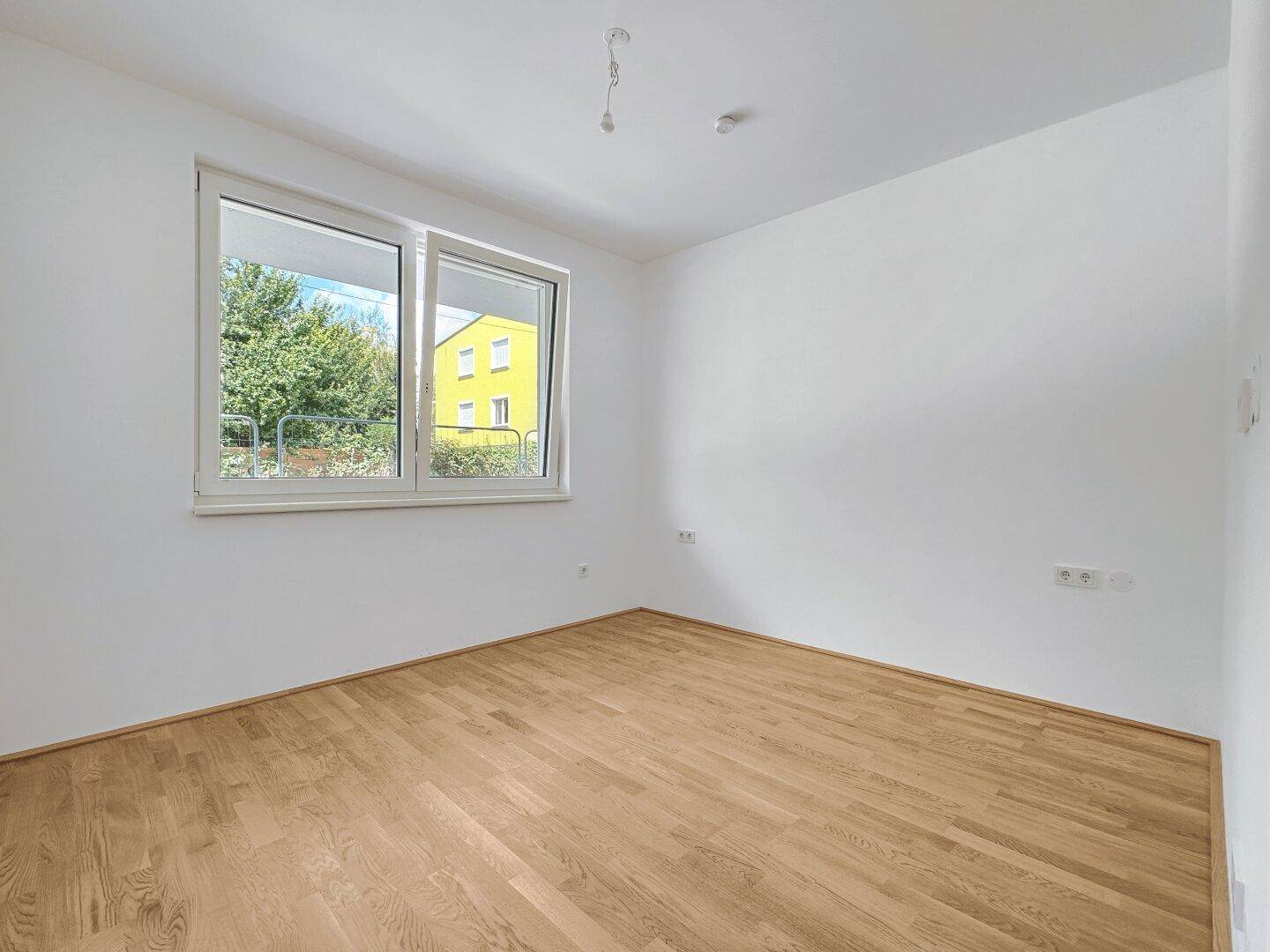 Heller Raum mit Parkettboden und großem Fenster, bietet Blick auf die umliegende Bebauung.