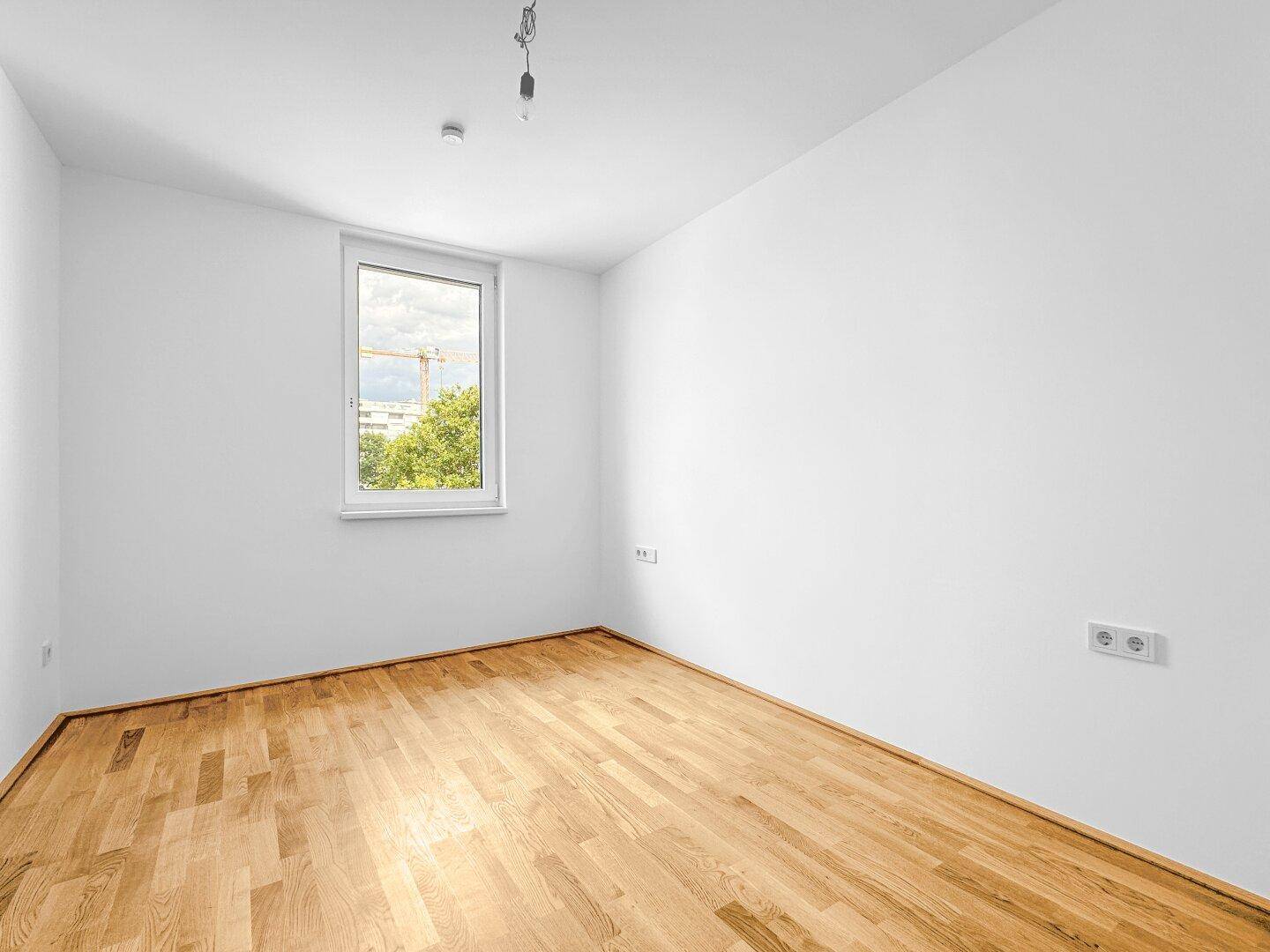 Helles Schlafzimmer mit Parkettboden und einem Fenster mit Blick ins Grüne und auf die Stadt.