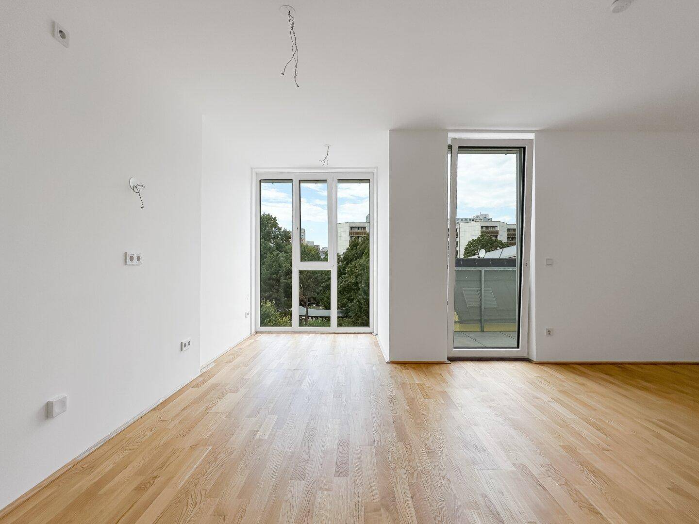 Helles, leeres Zimmer mit Holzboden und großen Fenstern, die einen Blick ins Grüne bieten.