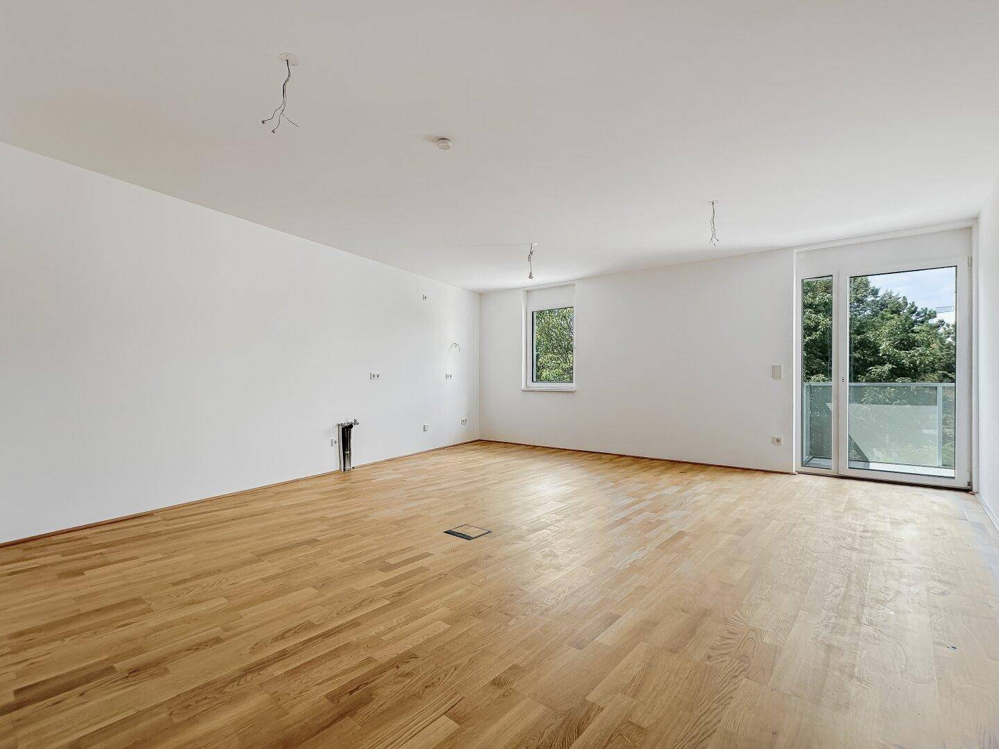 Geräumiger, heller Wohnraum mit Holzboden, Fenster und Zugang zu einem Balkon mit Grünblick.