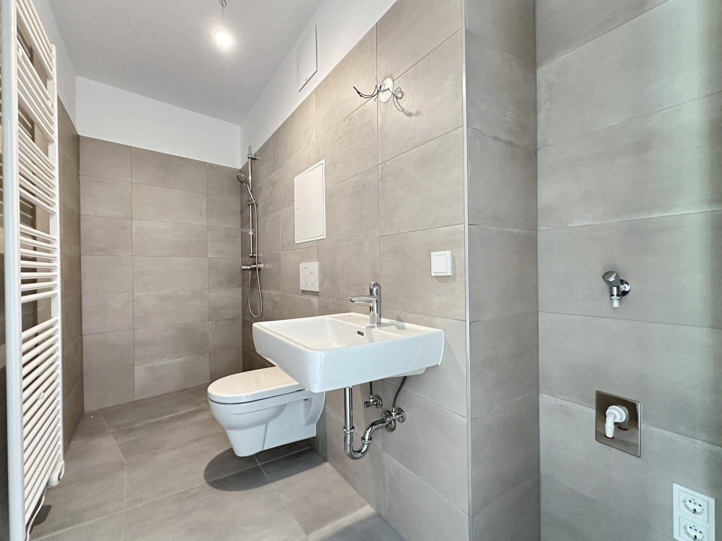 Modernes Badezimmer mit grauen Fliesen, wandhängendem WC, Waschbecken und Handtuchheizkörper.