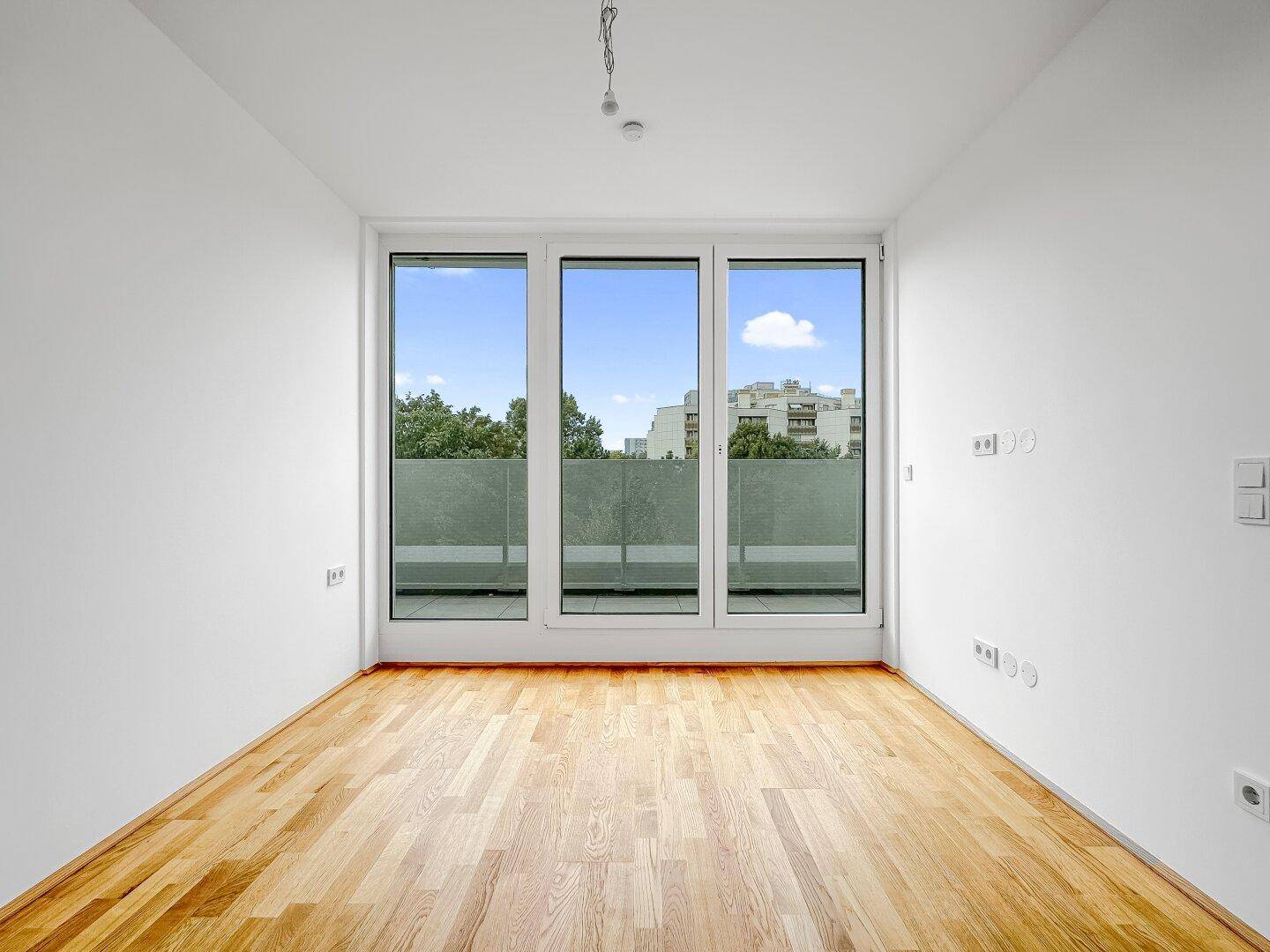 Helles Zimmer mit Holzboden und bodentiefen Fenstern, die einen weiten Blick ins Grüne ermöglichen.