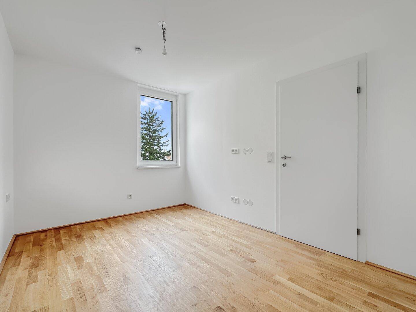Leeres Schlafzimmer mit hellem Parkettboden und einem Fenster, das einen Blick ins Grüne bietet.