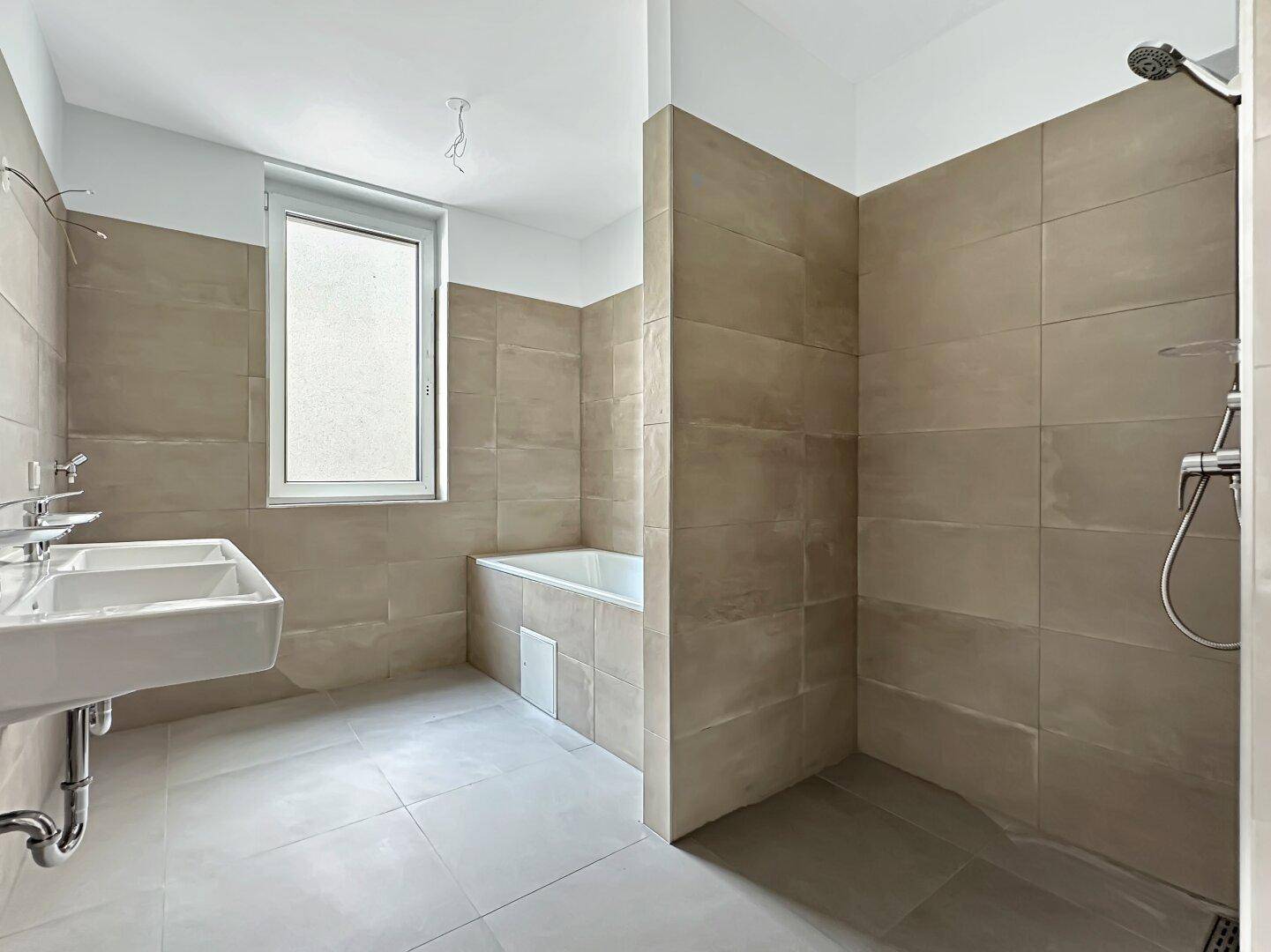 Modernes Badezimmer mit beigefarbenen Fliesen, Badewanne, Dusche und Fenster für natürliches Licht.