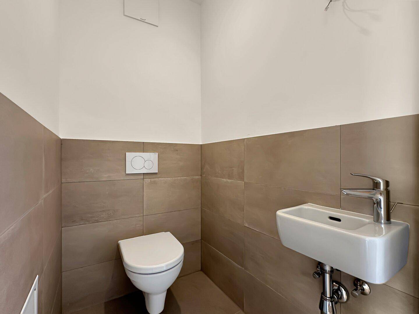 Separates WC mit Wand-WC, kleinem Waschbecken und beigefarbenen Wandfliesen.