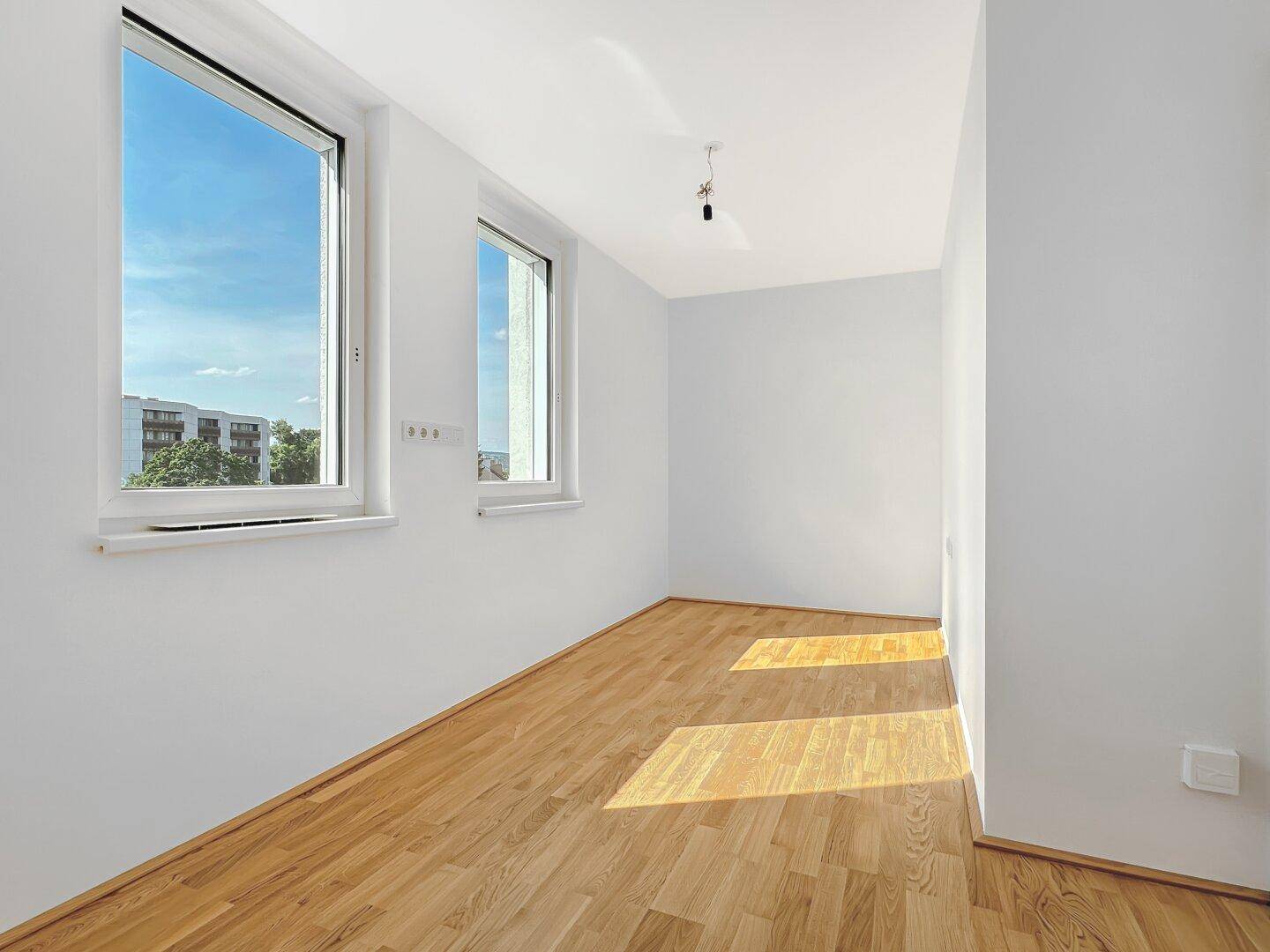 Helles, leeres Zimmer mit Holzboden und zwei Fenstern, die einen Blick ins Grüne bieten.
