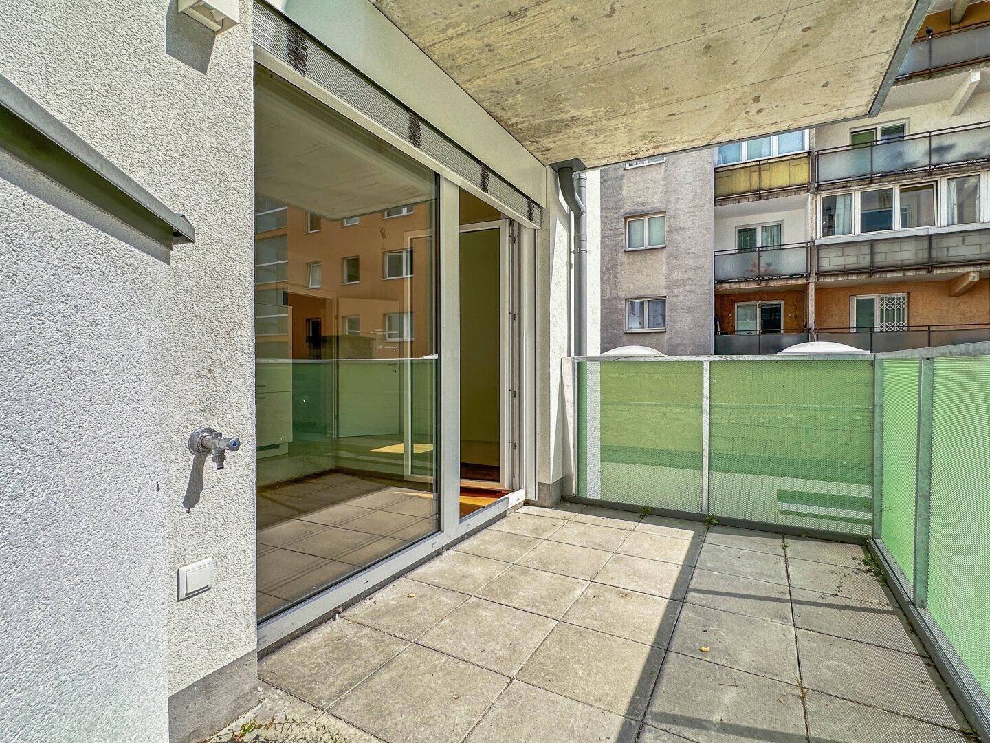 Heller Balkon mit Fliesenboden und grünen Sichtschutzelementen, umgeben von Gebäuden.