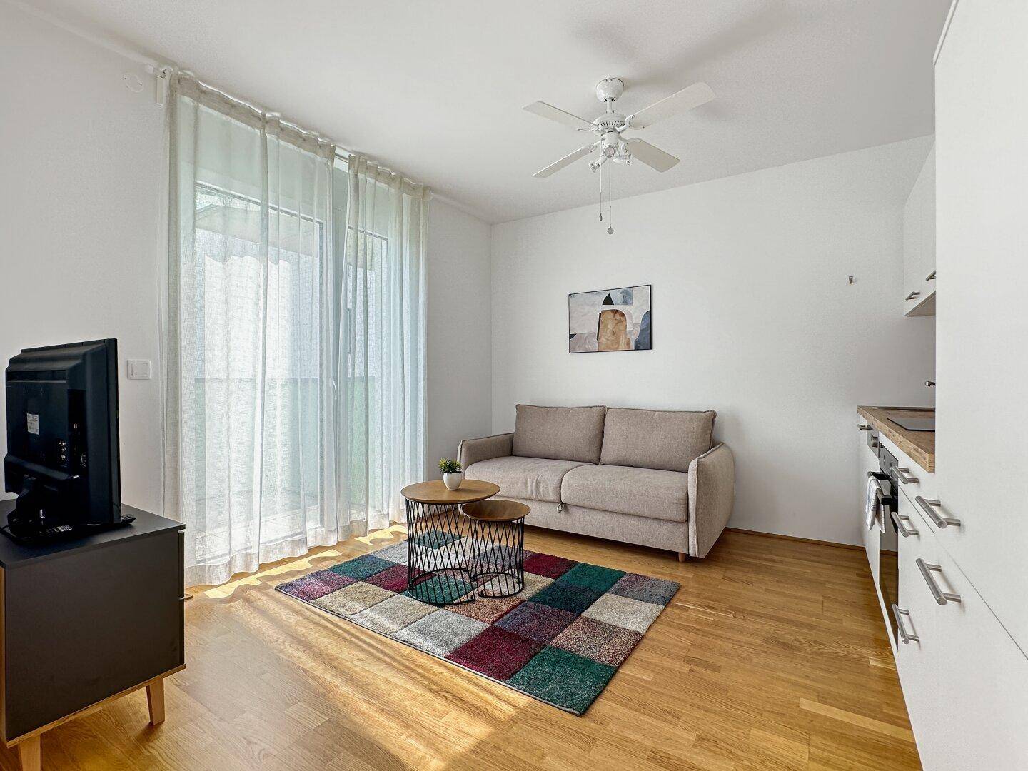 Gemütlicher Wohnbereich mit Sofa, Fernseher und Zugang zu einem Balkon durch große Fenster.