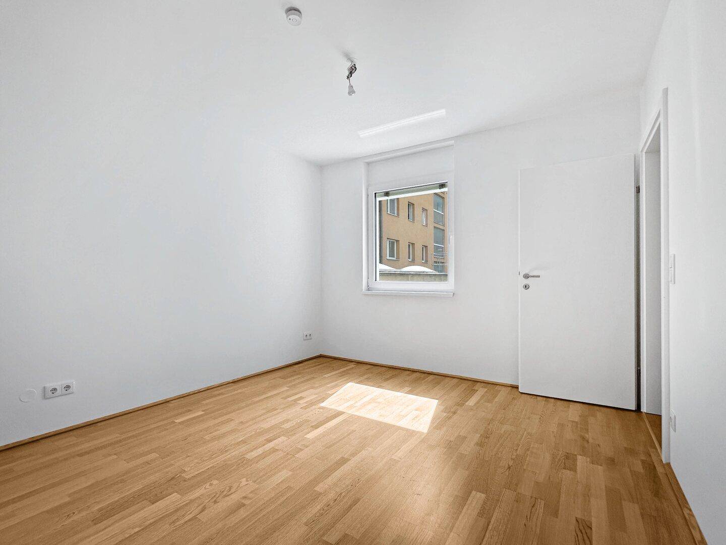 Leeres Zimmer mit hellem Parkettboden und einem Fenster, das Tageslicht hereinlässt.