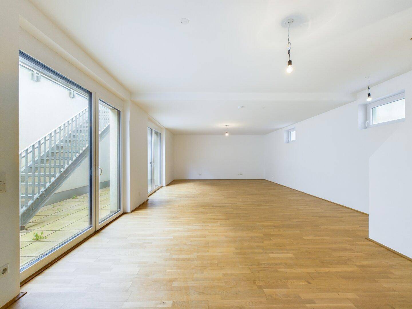 Heller Wohnraum mit Holzboden und breiten Fenstertüren, die zu einem Außenbereich führen.