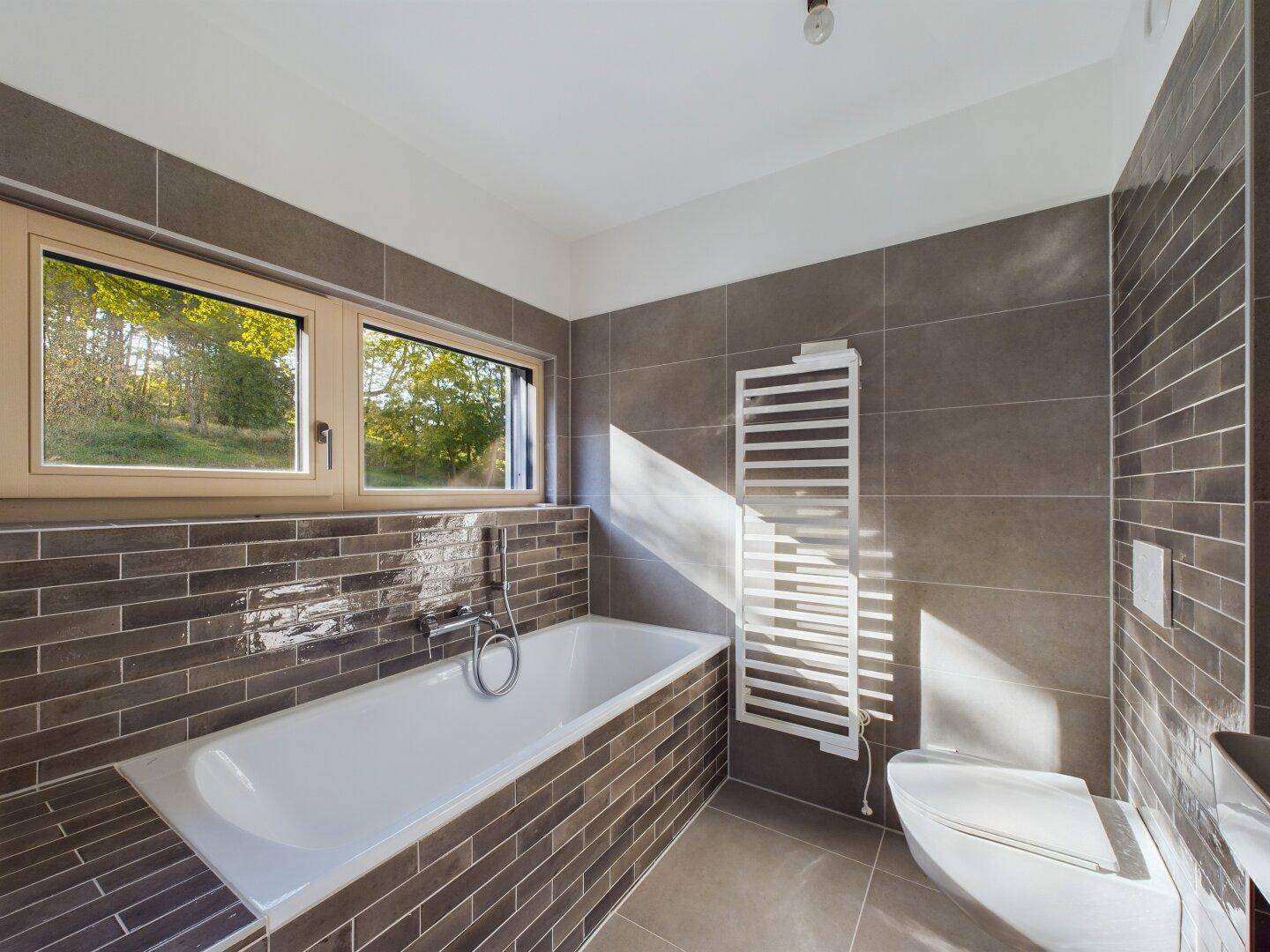 Modernes Badezimmer mit Badewanne, Wand-WC und beheiztem Handtuchhalter, gefliest in Grautönen.