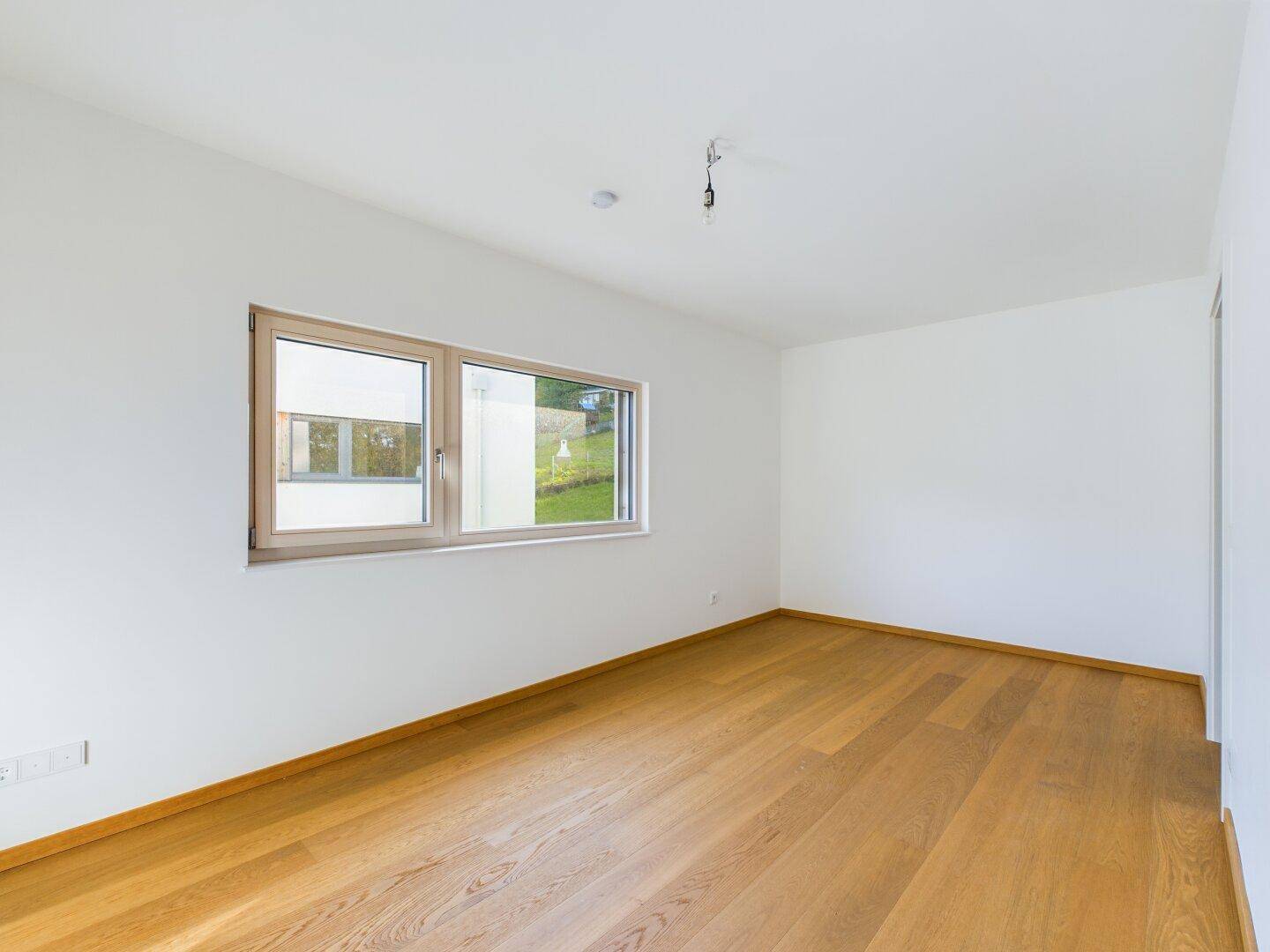 Helles Schlafzimmer mit Parkettboden und großem Fenster, das viel Tageslicht hereinlässt.