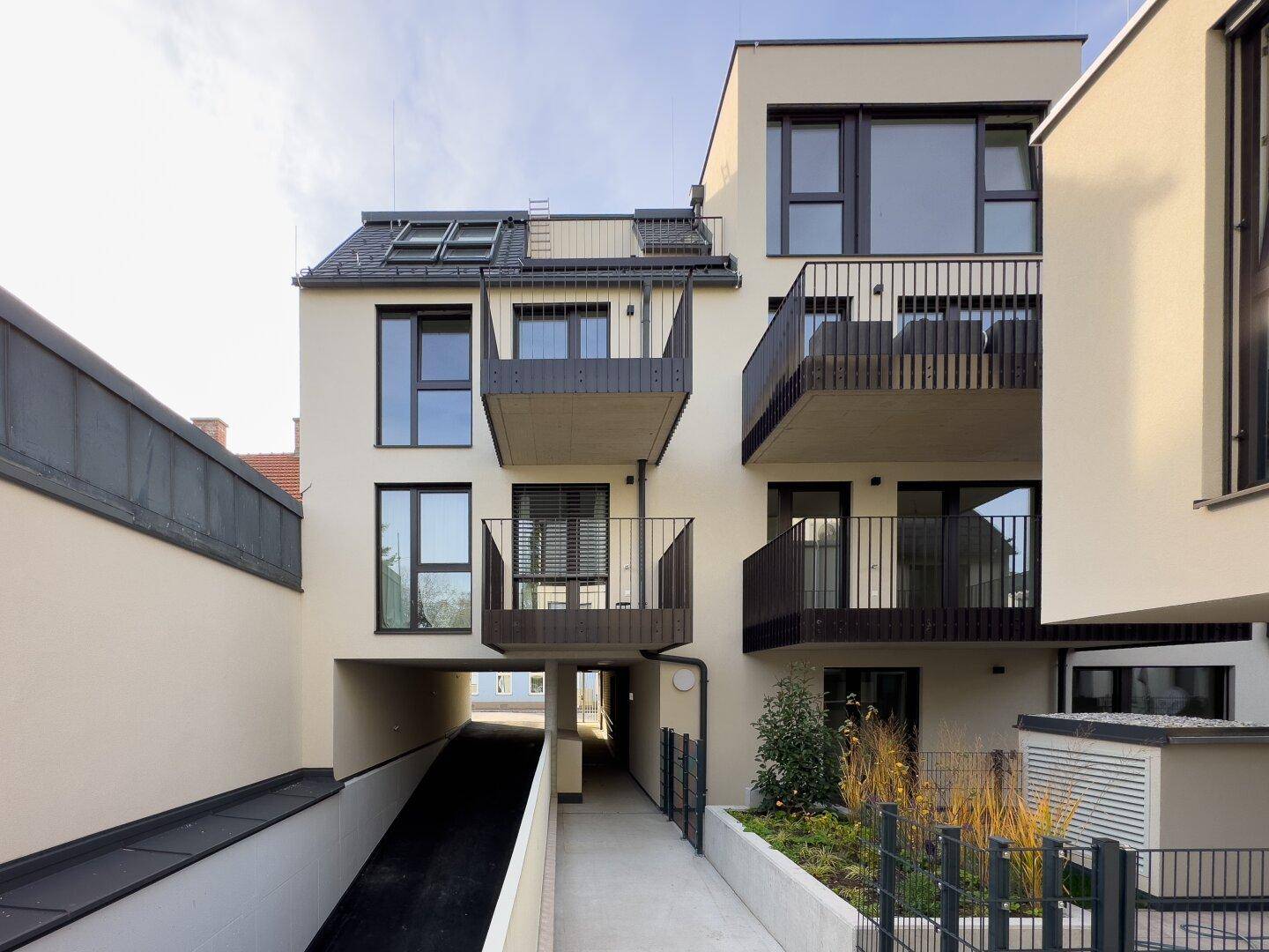 Rückansicht eines modernen Wohngebäudes mit Balkonen, Tiefgaragenzufahrt und begrüntem Eingangsbereich.