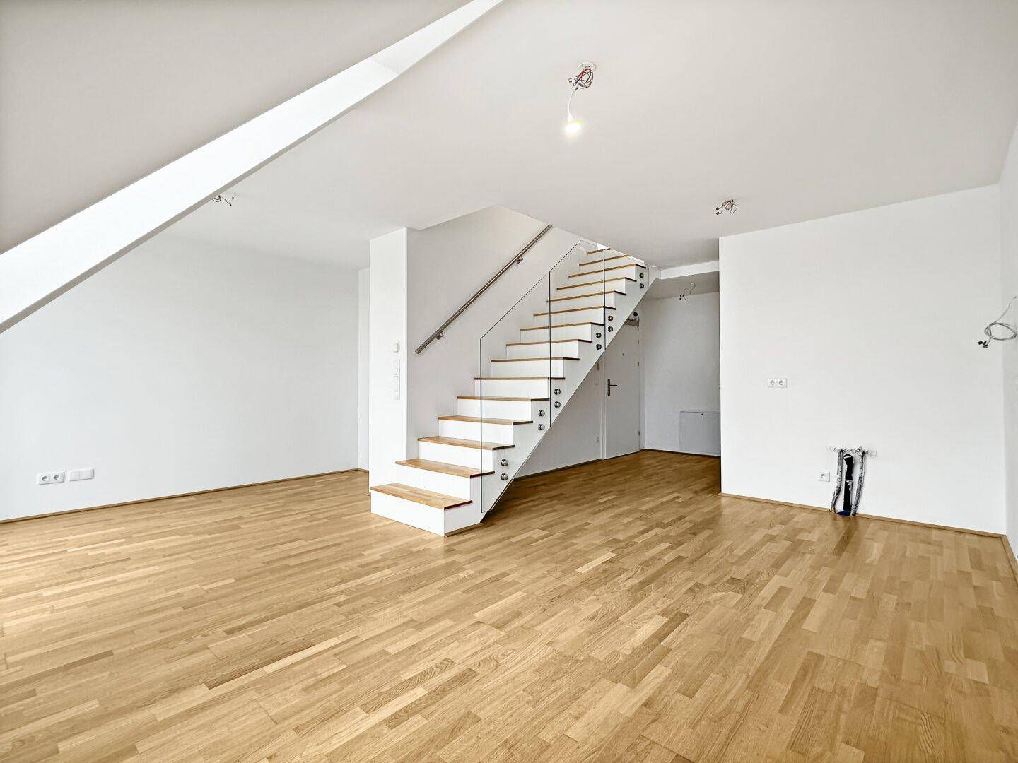 Offener Wohnbereich mit Holzparkett, moderner Treppe und viel Tageslicht durch große Fenster.
