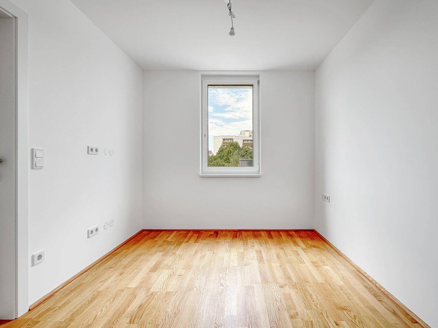 Leeres Zimmer mit hellem Parkettboden und einem Fenster mit Blick ins Grüne.