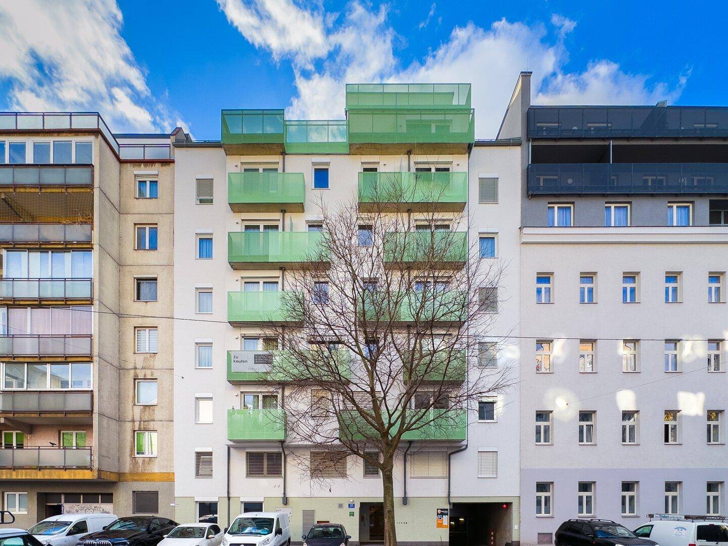 Mehrfamilienhaus mit moderner weißer Fassade, markanten grünen Balkonen und einem Baum davor.