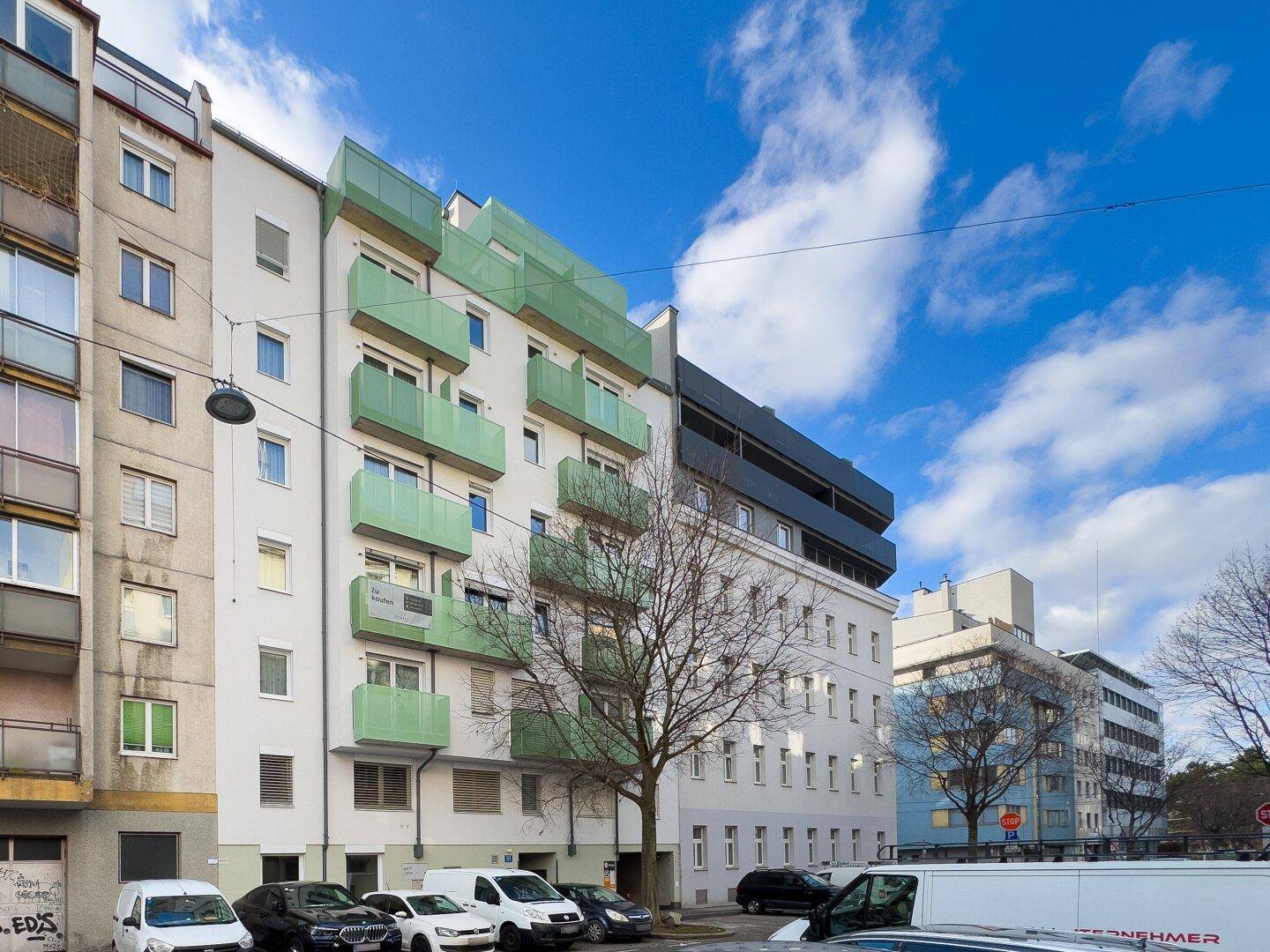 Städtisches Mehrfamilienhaus mit grünen Balkonen, umgeben von anderen Gebäuden in einer belebten Straße.