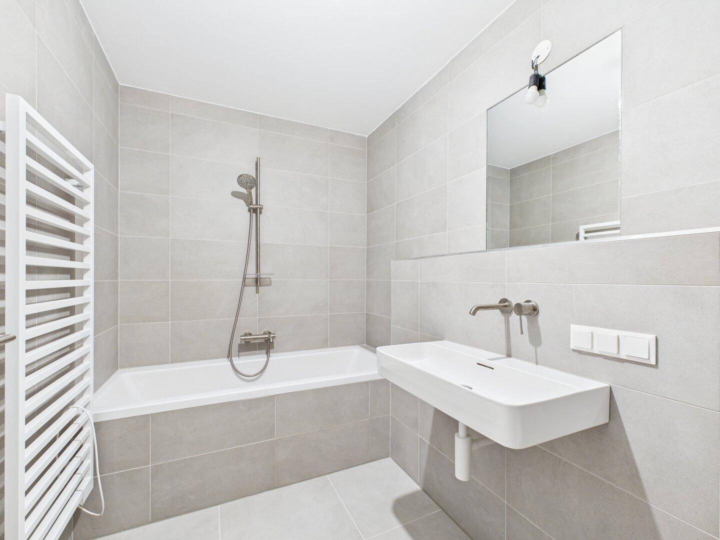Modernes Badezimmer mit hellgrauen Fliesen, Badewanne und praktischem beheiztem Handtuchhalter.