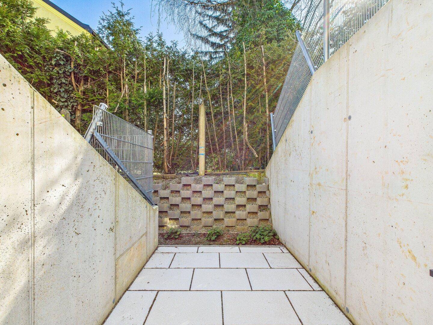 Schmaler gepflasterter Weg zwischen Betonwänden, der zu einem begrünten Bereich führt.