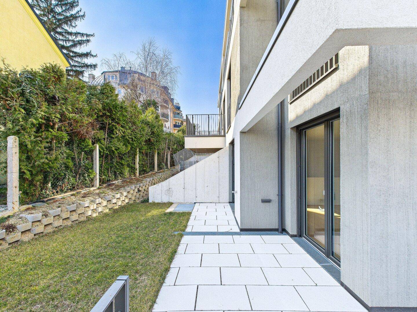 Seitlicher Gartenbereich mit gepflastertem Weg, Rasenfläche und grüner Heckenbepflanzung.