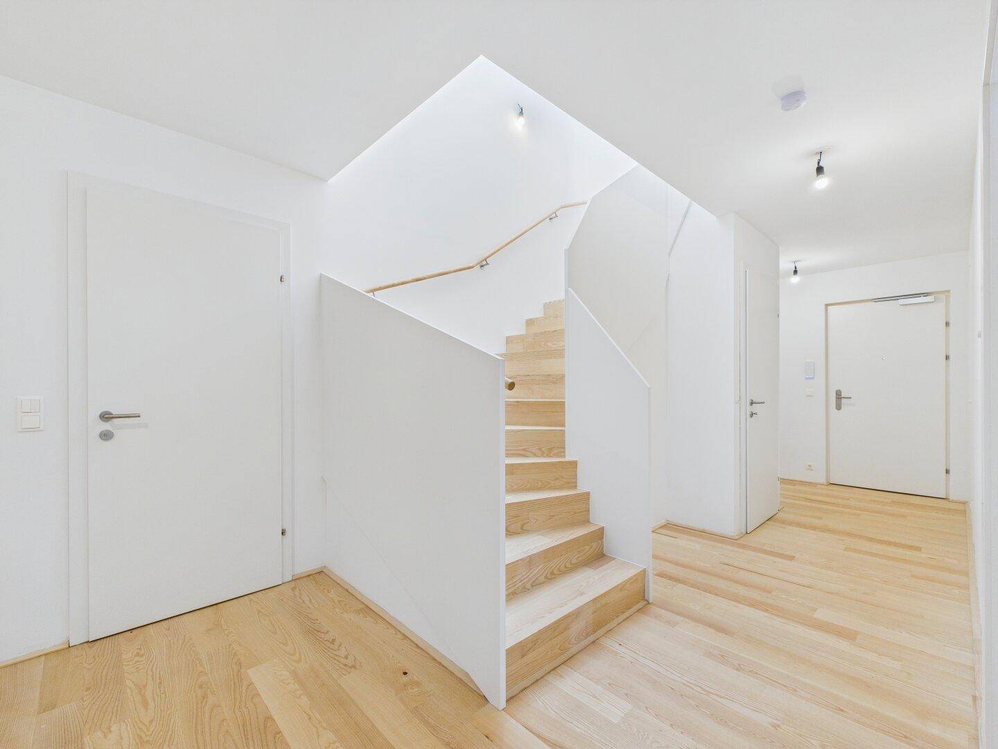Heller Flur mit moderner Holztreppe, weißen Wänden und natürlichem Lichteinfall von oben.