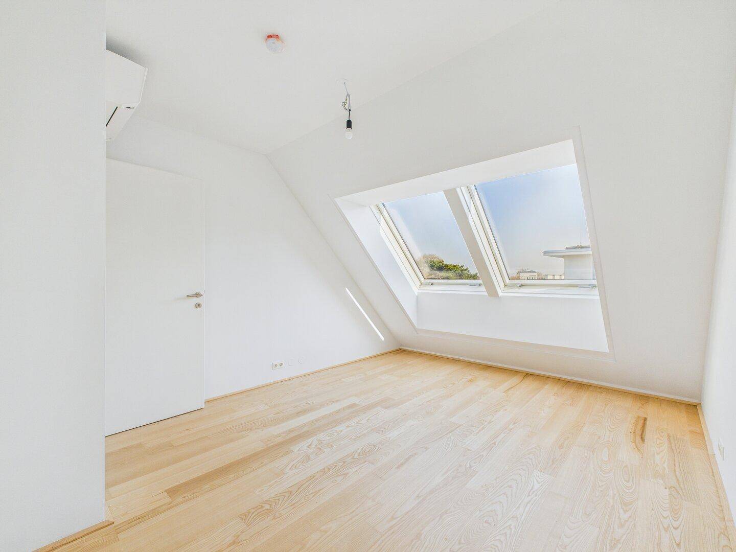 Lichtdurchflutetes Zimmer im Dachgeschoss mit Schrägfenster und Klimaanlage an der Wand.