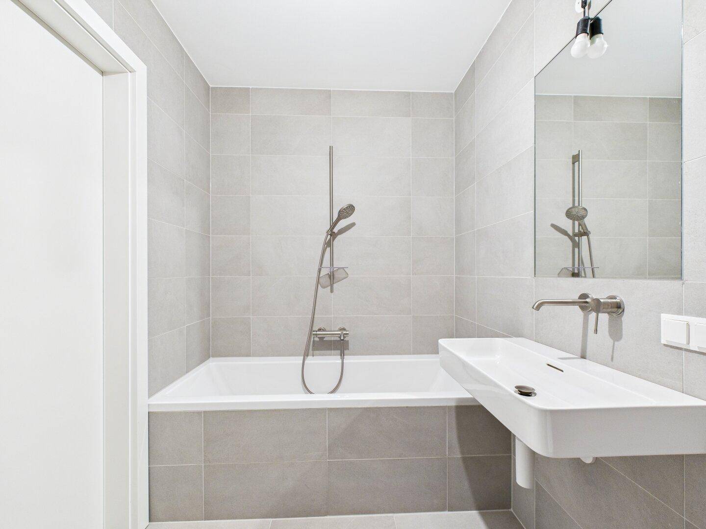 Modernes Badezimmer mit hellgrauen Fliesen, Badewanne und einem schwebenden Waschbecken.