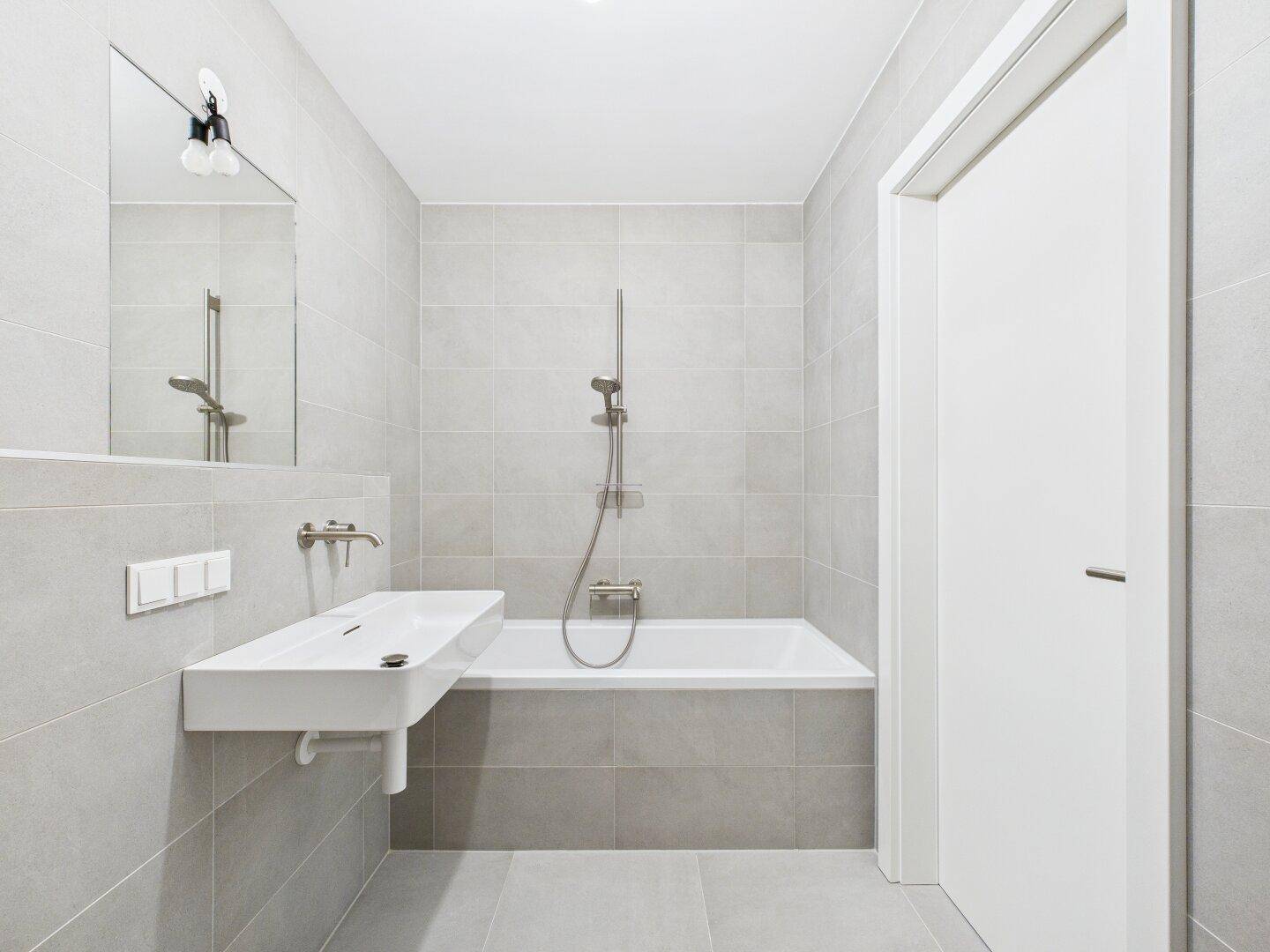 Zeitgemäßes Badezimmer mit Badewanne, Waschtisch und großem Spiegel, in neutralen Grautönen gehalten.