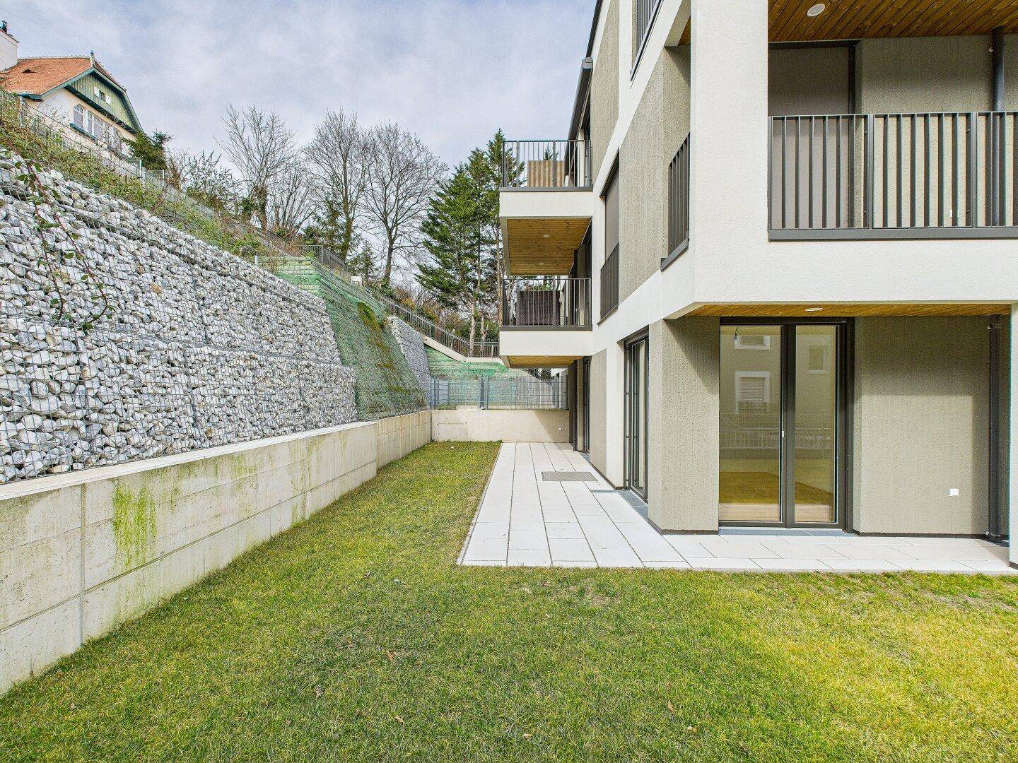 Außenansicht des Gebäudes mit Terrasse, Rasenfläche und einer stabilen Gabionenwand im Hintergrund.