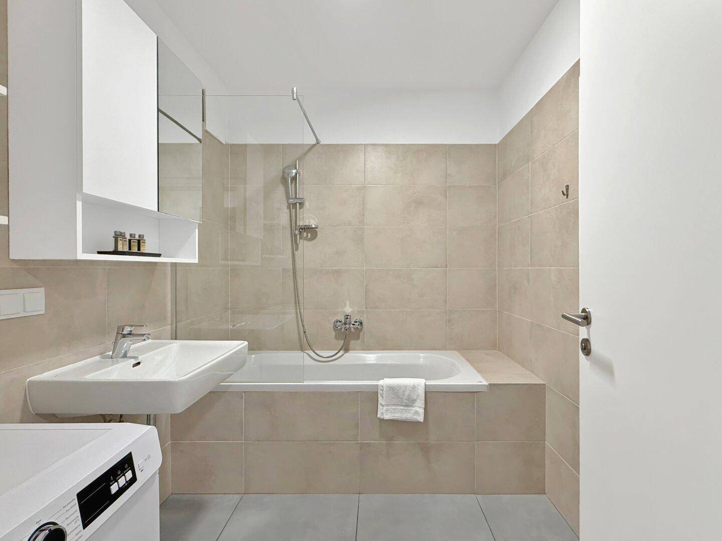 Badezimmer mit Badewanne, Waschbecken und hellen Fliesen an Wänden und Boden.