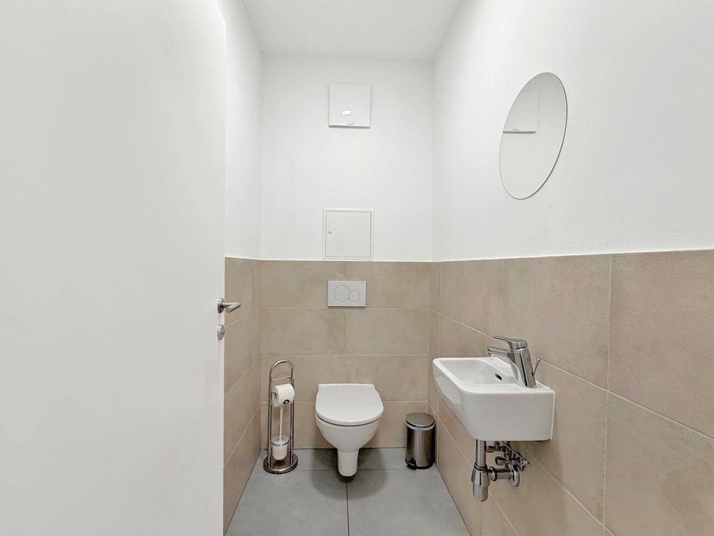 Separates WC mit wandhängendem WC, kleinem Waschbecken und modernen Fliesen.