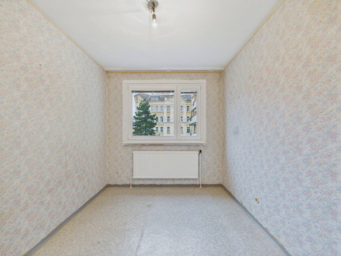 Helles Zimmer mit Tapeten, Fensterblick ins Grüne und auf ein Gebäude.