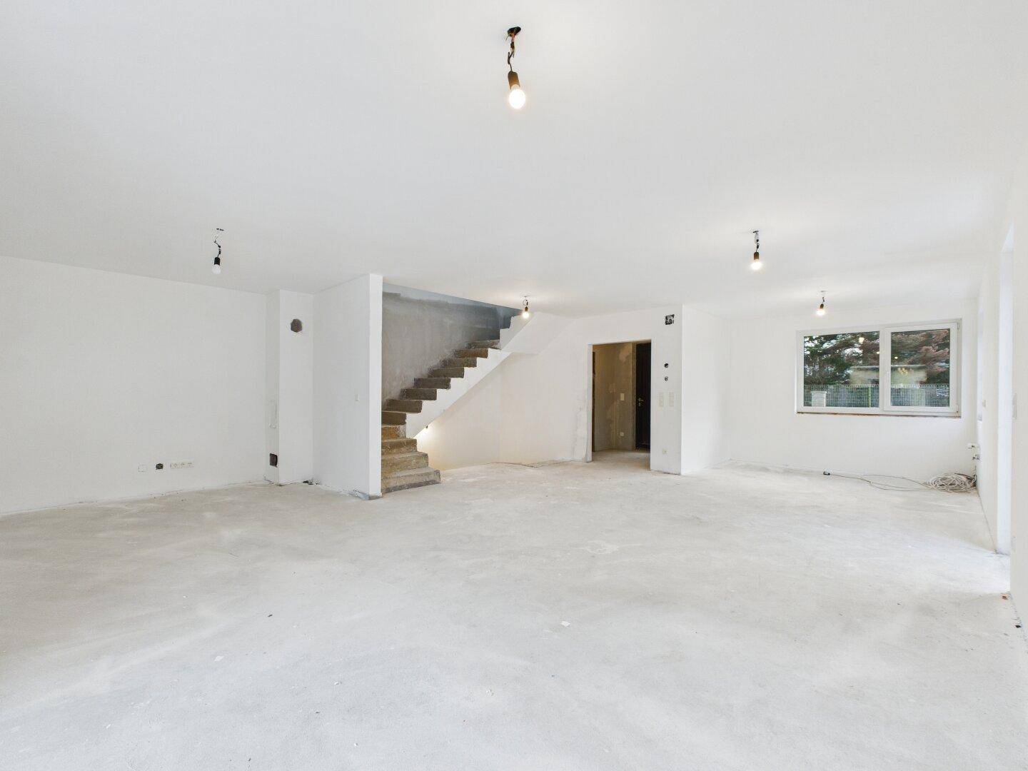 Großer, heller Raum im Rohbauzustand mit unfertiger Treppe und Fenstern, bereit für den Ausbau.