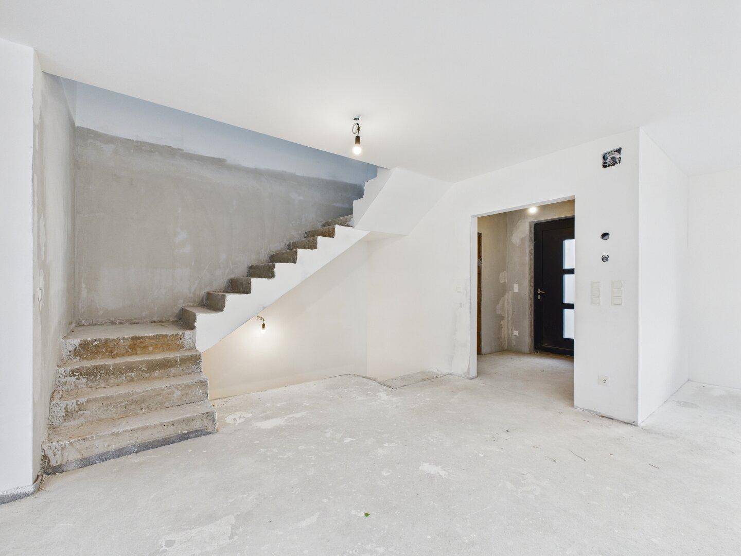 Eingangsbereich im Rohbau mit unfertiger Treppe und Blick auf die Haustür, bereit für den Innenausbau.