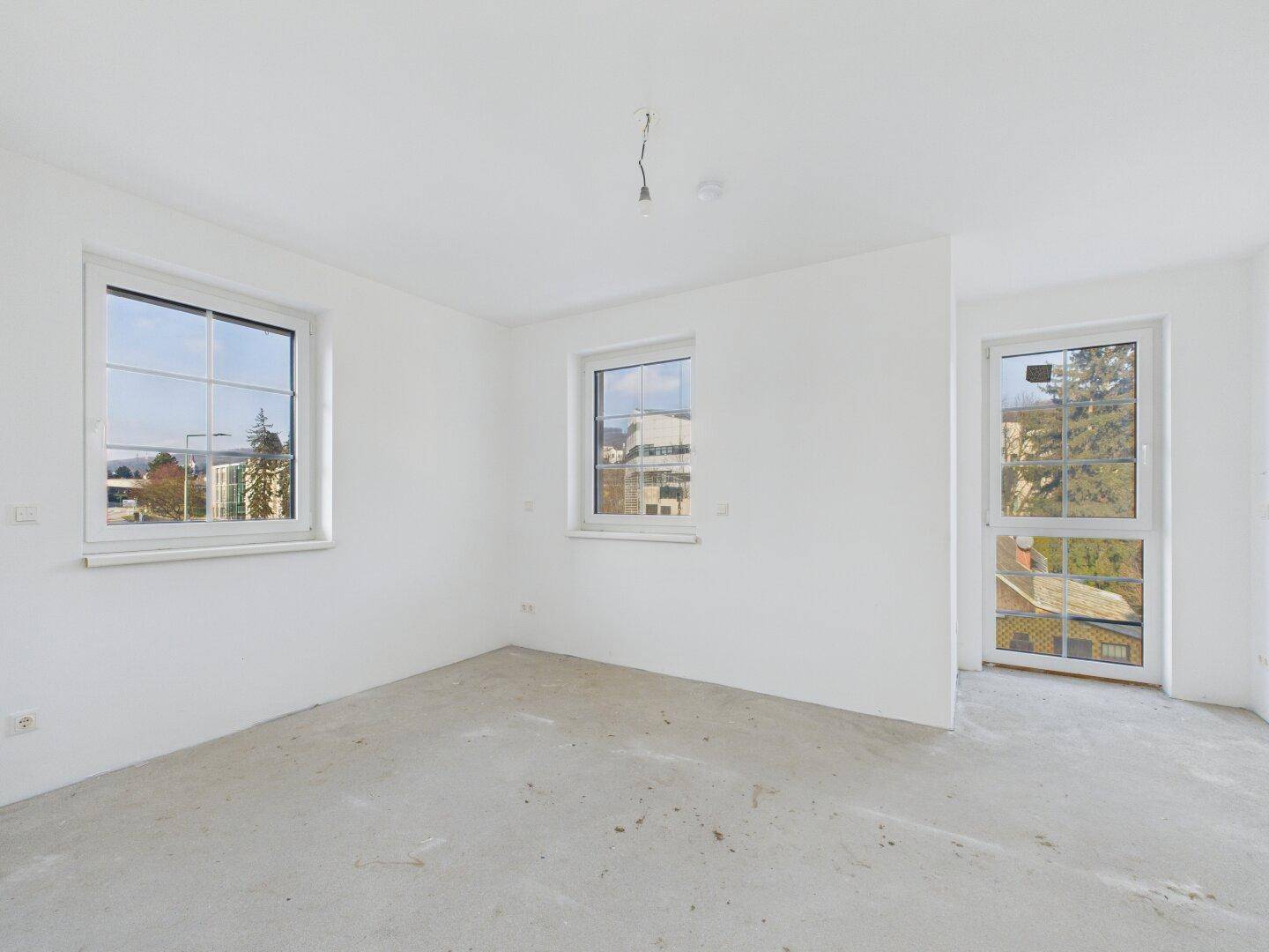 Heller Raum im Rohbau mit mehreren Fenstern und Zugang zu einem französischen Balkon.