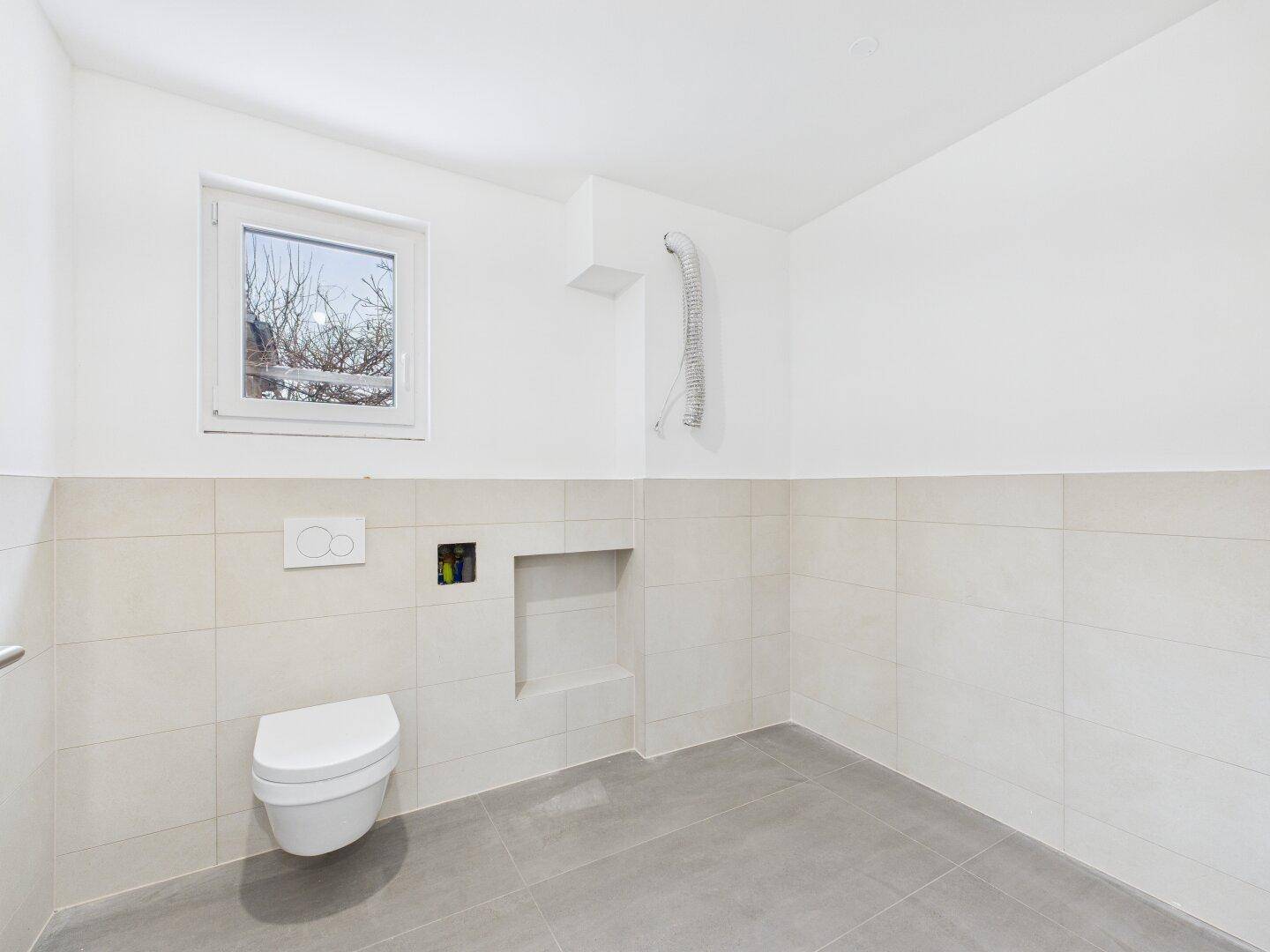 Modernes Gäste-WC mit wandhängender Toilette, Fenster und hellen Fliesen, bereit zur Nutzung.