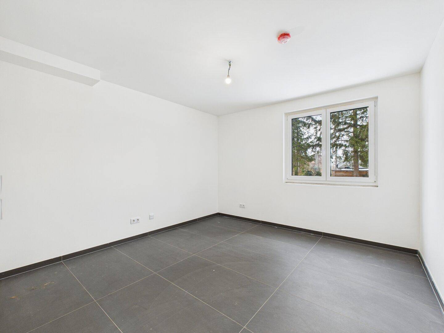 Helles Zimmer mit großem Fenster und dunklen Fliesen, ideal als Schlafzimmer oder Arbeitszimmer.
