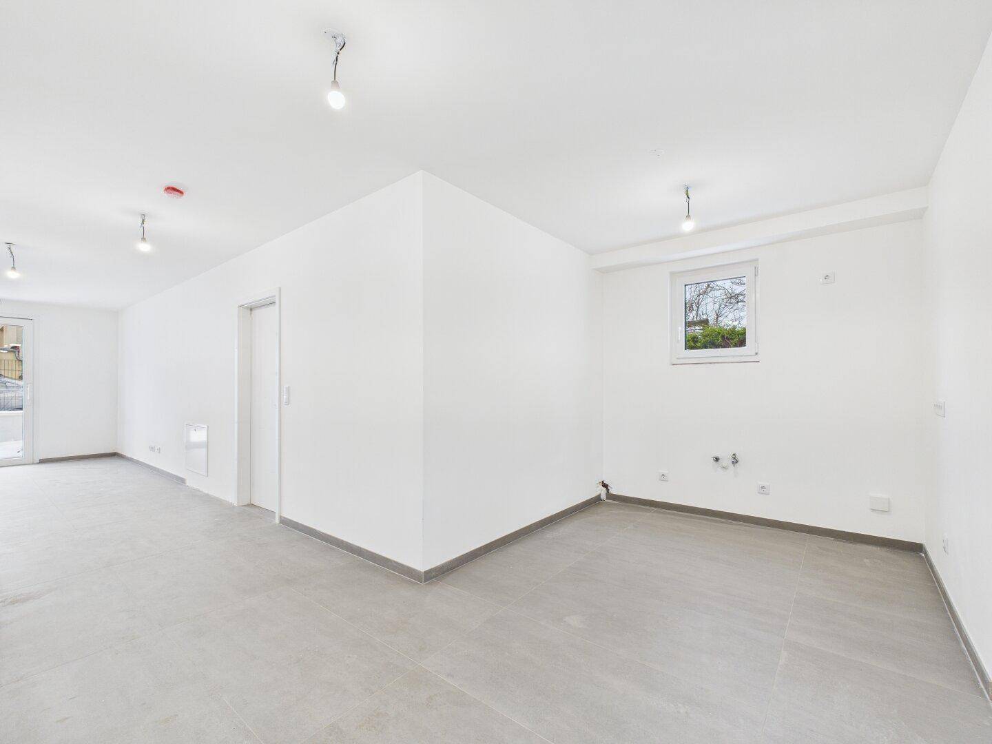 Offener Wohnbereich mit hellen Wänden und Fliesenboden, vorbereitet für Kücheninstallationen in der Ecke.