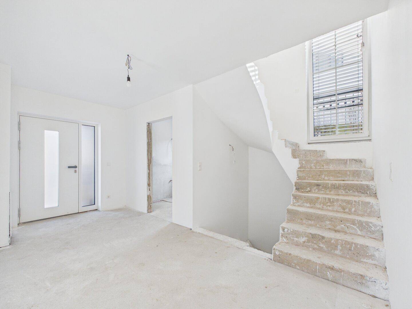 Eingangsbereich im Rohbau mit Treppe zum Obergeschoss und viel natürlichem Licht durch das große Fenster.